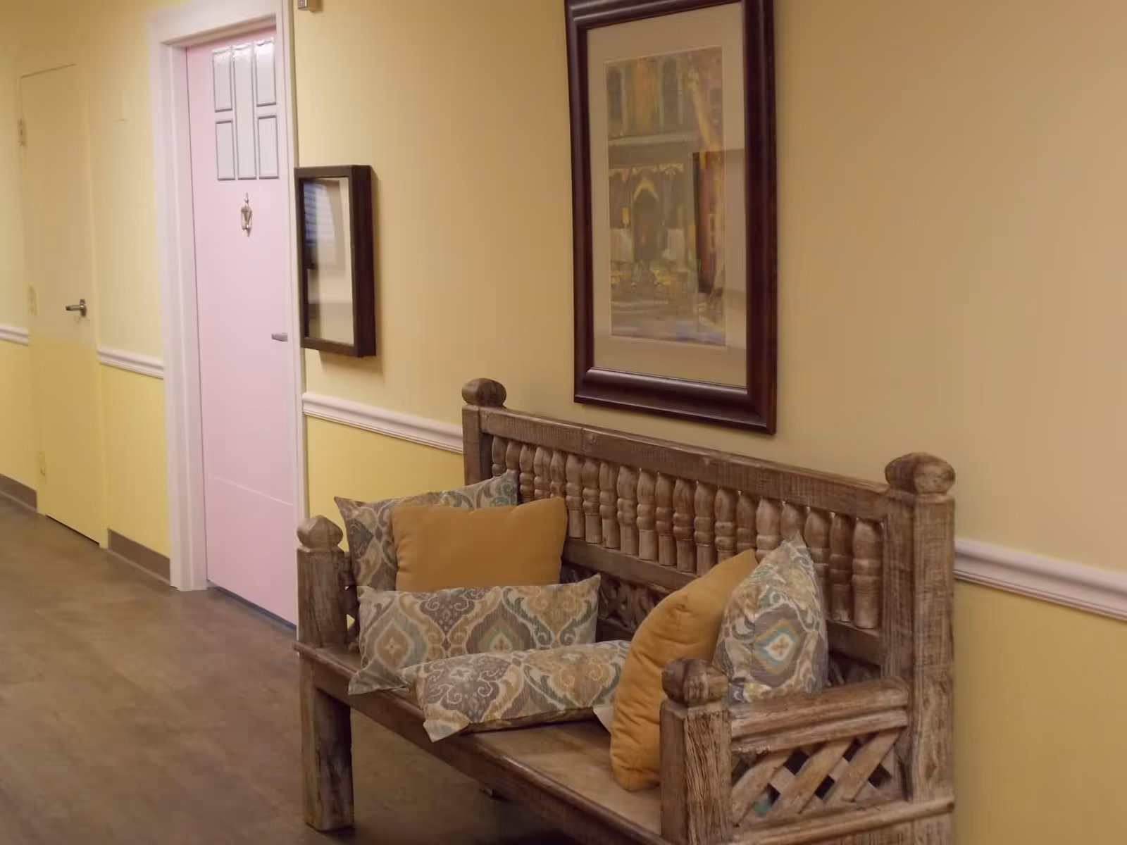 A hallway with yellow walls and wooden flooring featuring a rustic wooden bench with patterned and solid-colored cushions. There is a framed picture hanging on the wall above the bench and a pink door with decorative panels on the left side.