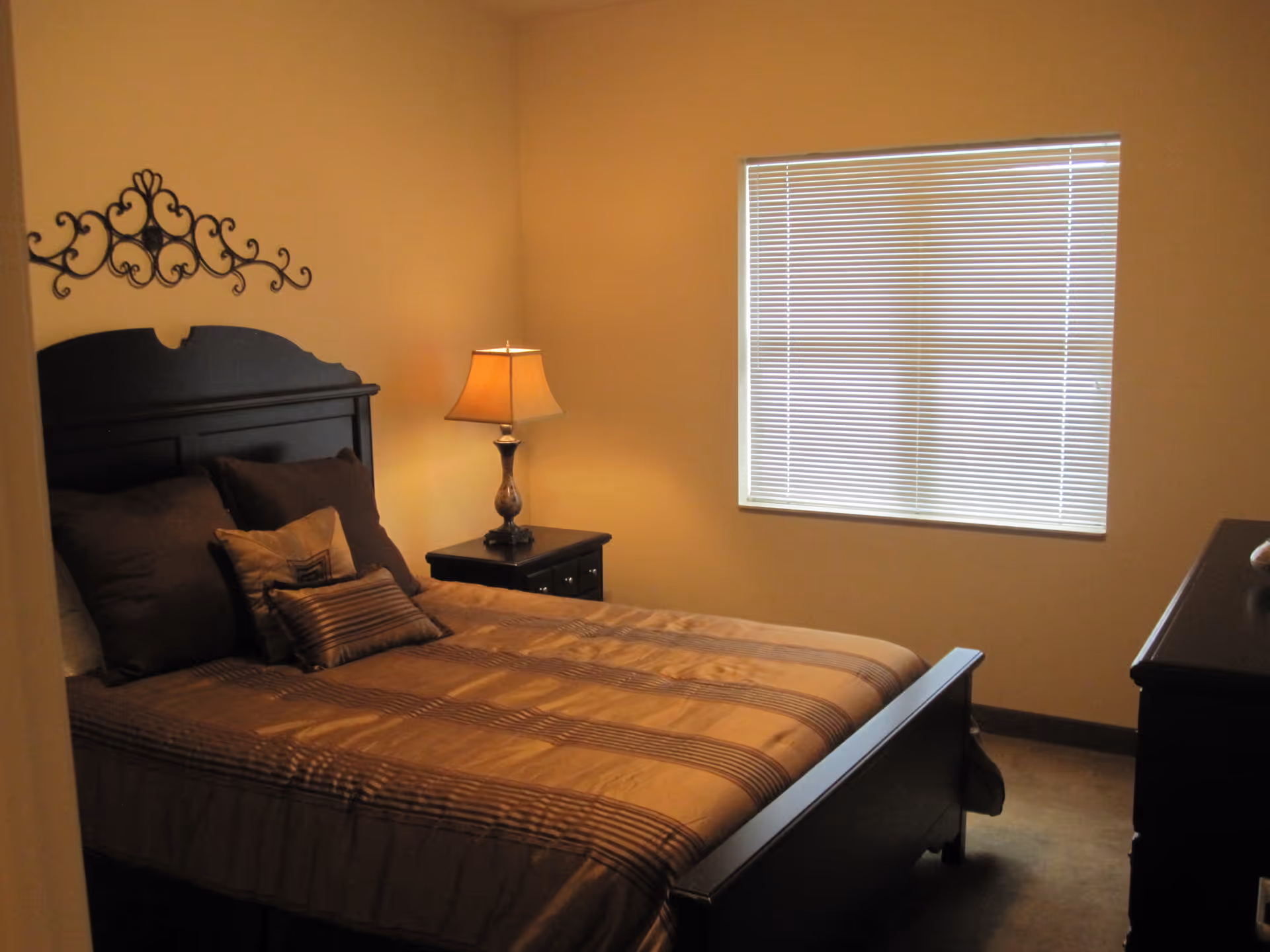 A bedroom with a dark wooden bed frame and matching nightstand. The bed is made with brown and beige striped bedding and several pillows. A lamp with a beige shade is on the nightstand. There is a window with closed blinds on the wall opposite the bed, and a decorative metal wall hanging above the headboard.