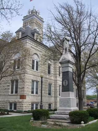 Stone historic building with a statue monument, flag, and leafless trees on a grassy lawn.