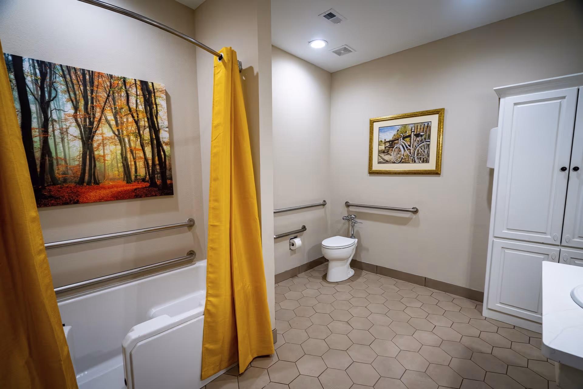 A spacious bathroom with beige walls and hexagonal floor tiles. The room features a white toilet with grab bars on the walls around it, a bathtub with a yellow shower curtain, and a white cabinet. There are two framed pictures on the walls, one depicting a forest scene and the other a bicycle by a fence.