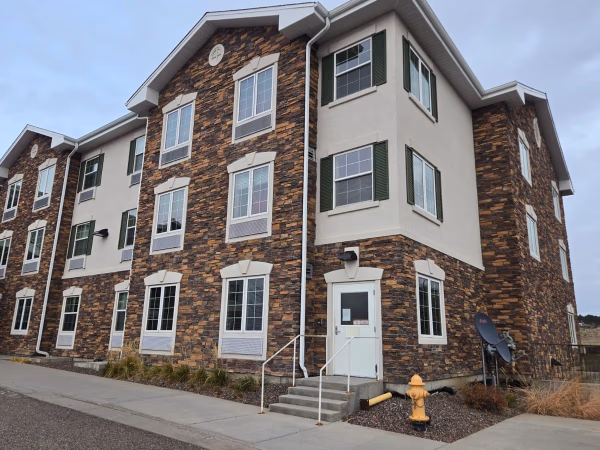 Three-story stone-and-stucco senior living building with multiple windows, a white entry door accessed by steps, and a yellow fire hydrant in front.
