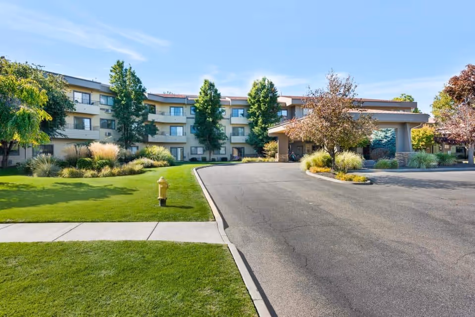 Exterior view of a multi-story senior living facility named Royal Columbian with a driveway, green lawn, trees, and shrubs under a clear blue sky.