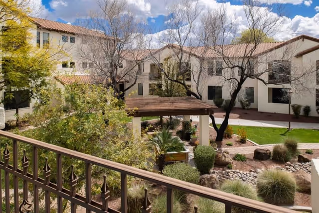 View of an outdoor courtyard area at Villa Hermosa with a wooden pergola, various trees and shrubs, rocks, and a well-maintained lawn surrounded by a two-story building with beige walls and red tile roofing under a partly cloudy sky.