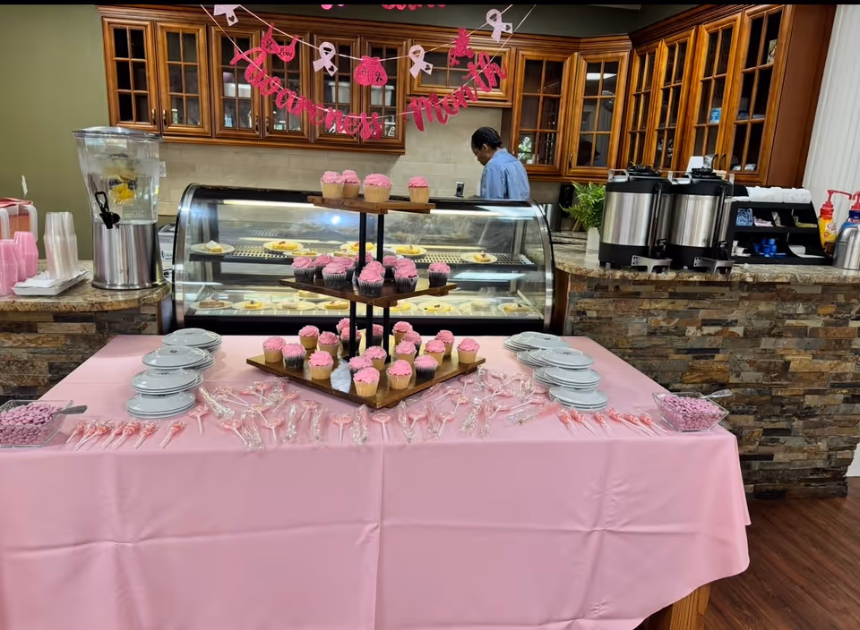 Pink-themed dessert table with cupcakes, lollipops, plates and a drink dispenser set up in front of a refrigerated display case and coffee urns in a communal dining area.