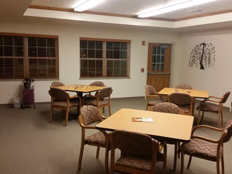 A well-lit room with several square tables and cushioned chairs arranged around them. The room has beige walls, two windows with wooden blinds, a door with a window, and a decorative tree wall art. There is a small cabinet with a plant on top near the windows.
