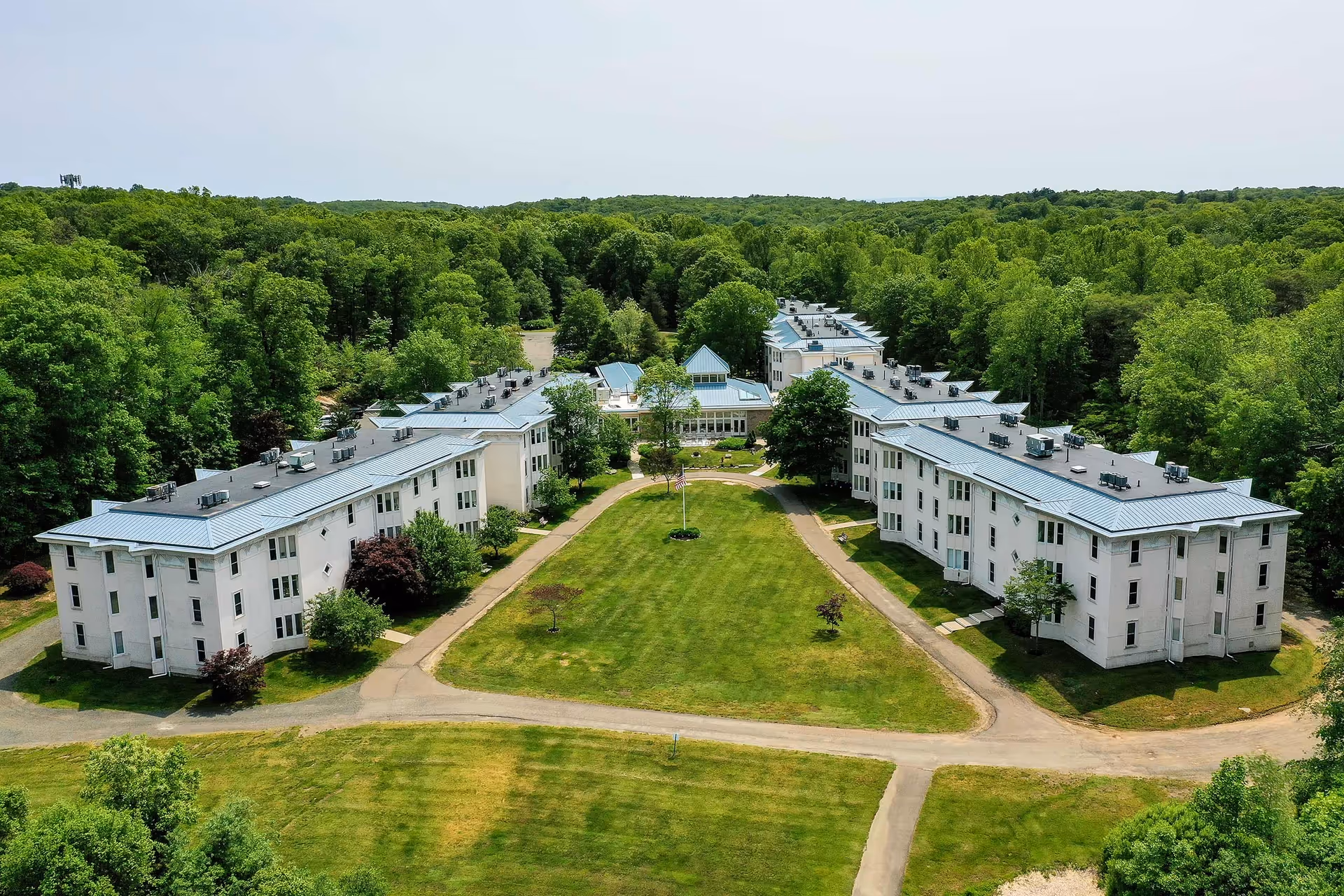 Aerial view of Guilford Retirement Residence showing two large white buildings with blue-gray roofs facing each other across a green lawn with a flagpole in the center. The buildings are surrounded by dense green trees and pathways.