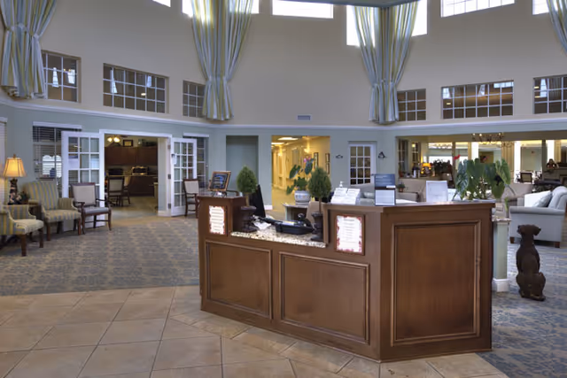 Reception area of an assisted living facility with a wooden front desk, potted plants, and seating areas with armchairs. The space has high ceilings with large windows and striped curtains, and there are doorways leading to other rooms including a kitchen area.