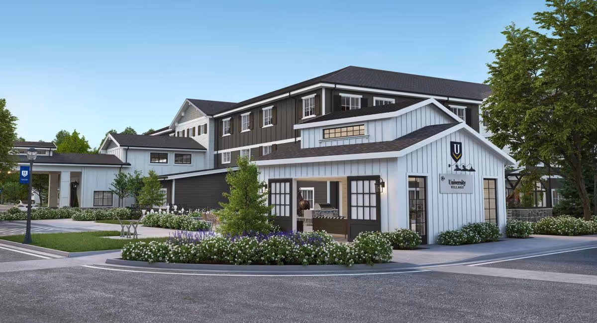 Exterior view of a modern senior living facility building with white and dark gray siding, multiple windows, and a landscaped area with bushes and trees. The building has a sign that reads 'University Village' and a logo with the letter U. The sky is clear and blue.