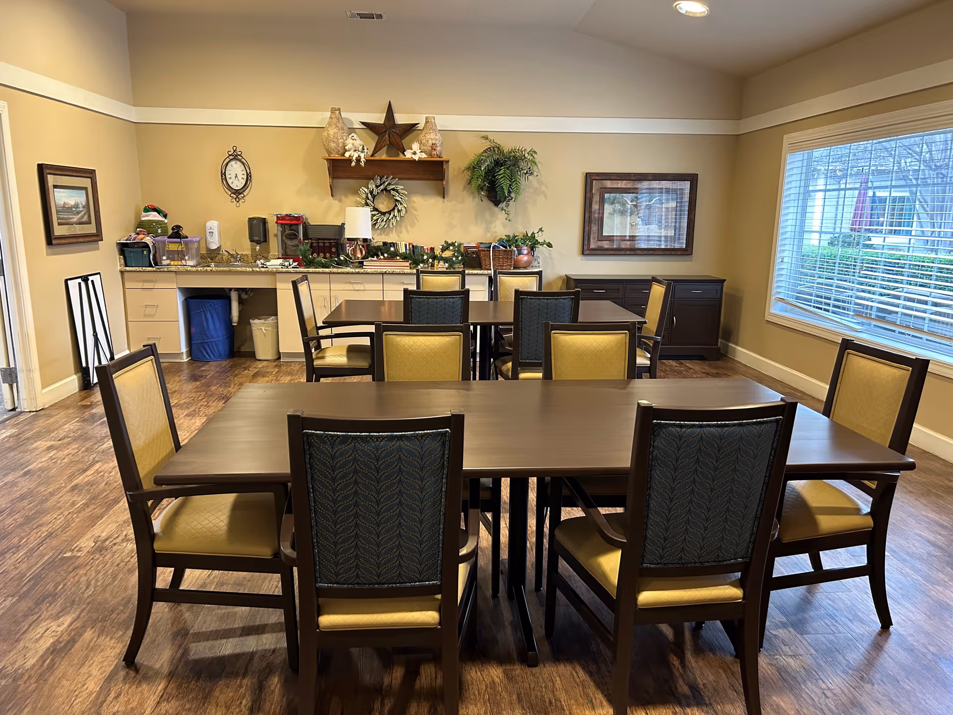 A dining room with two rectangular wooden tables and several chairs with yellow and blue upholstery. The room has wooden flooring, beige walls, and a large window with white blinds letting in natural light. On the back wall, there is a counter with various items including a coffee maker, decorations, and plants. A framed picture and a clock are also visible on the walls.
