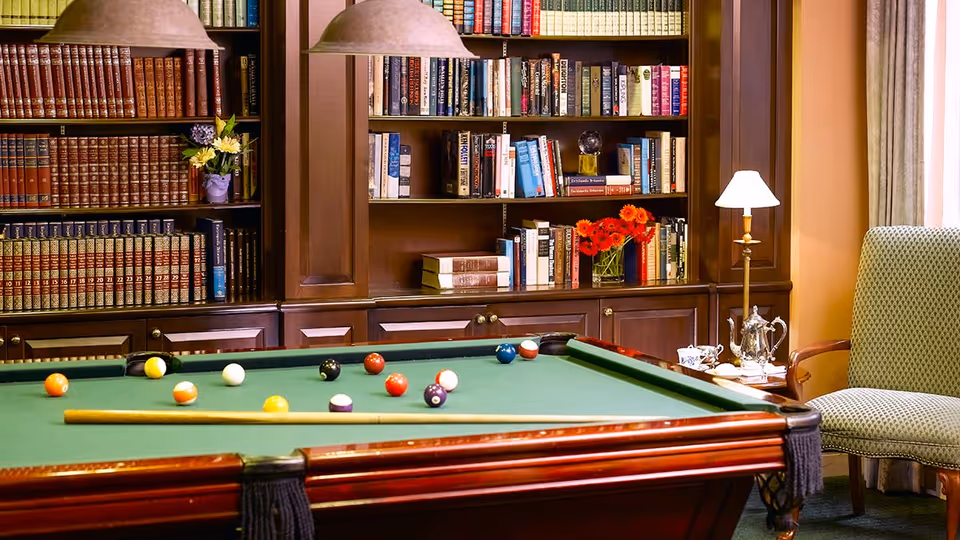 A cozy interior room featuring a green pool table with billiard balls and a cue stick on it. Behind the pool table is a large wooden bookshelf filled with books and decorative items, including flowers in vases. To the right, there is a cushioned armchair next to a small table with a lamp and a silver tea set. The room has warm lighting and a comfortable, inviting atmosphere.