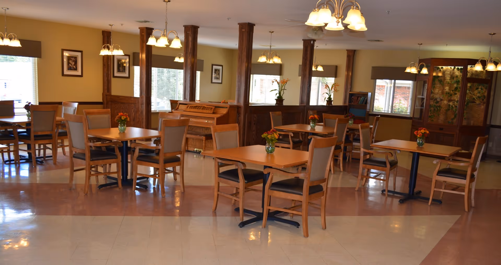 A dining room with multiple square wooden tables and chairs arranged neatly. Each table has a small vase with flowers. The room has large windows with blinds, wooden pillars, framed pictures on the walls, and several ceiling light fixtures. There is a piano against the far wall and a wooden cabinet with plants inside.