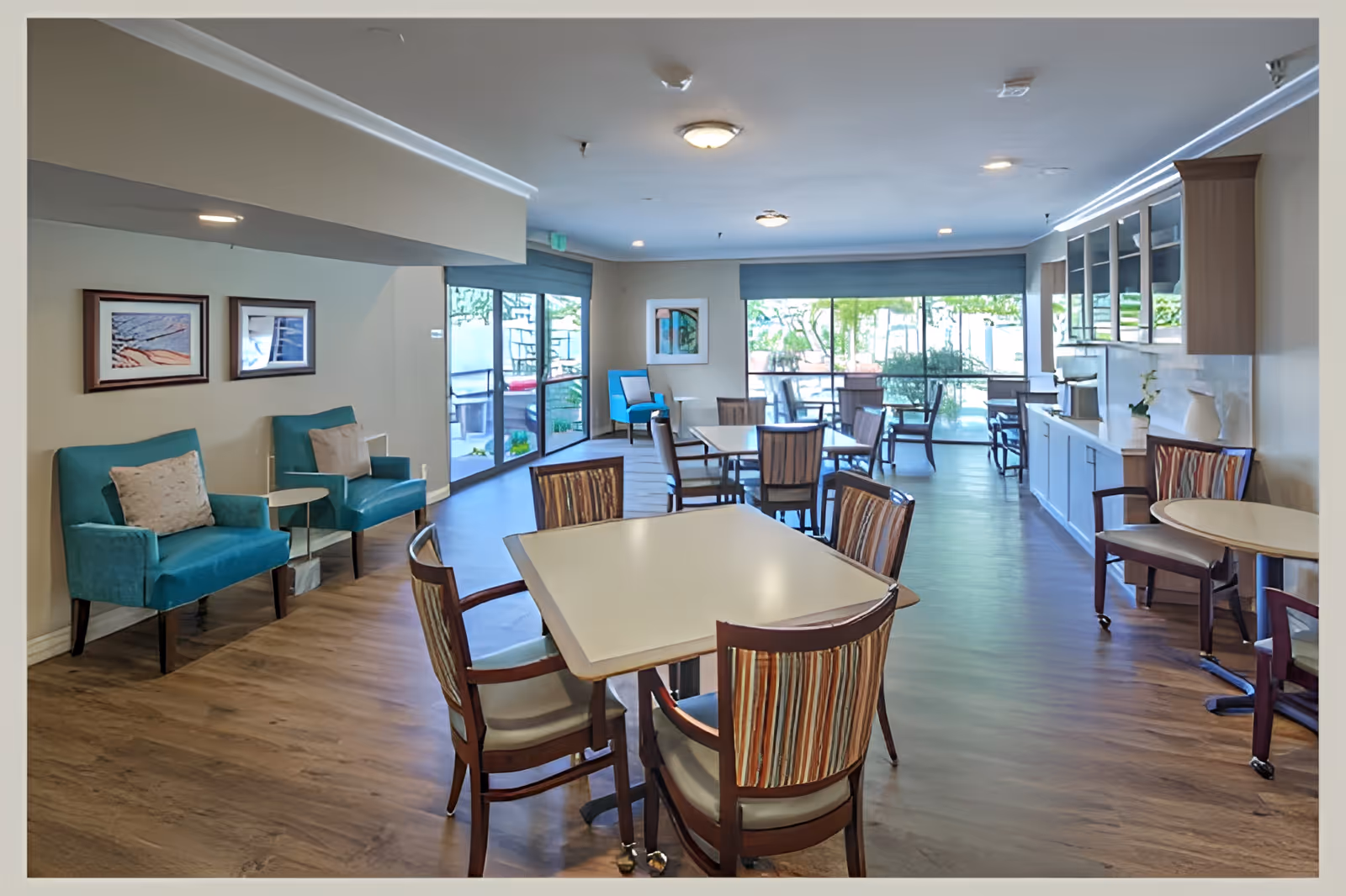 A bright and spacious senior living dining area with multiple tables and chairs arranged neatly. The room features large windows allowing natural light to fill the space, teal armchairs with cushions along one wall, framed artwork, and a built-in cabinet with a countertop on the right side. The flooring is wood-style, and the overall atmosphere is clean and welcoming.