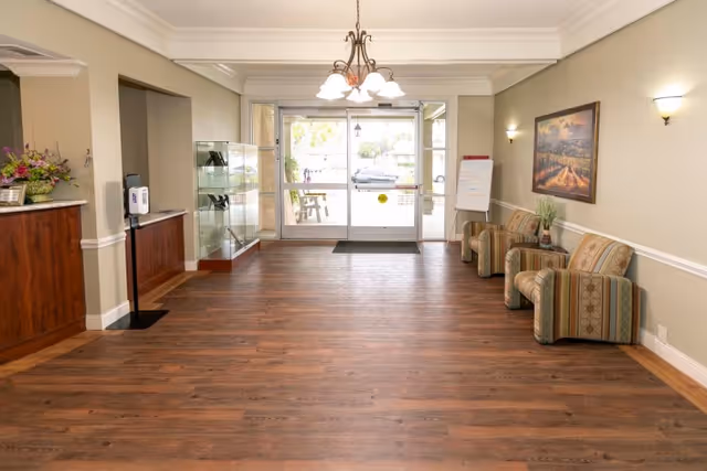 A spacious facility lobby with wooden flooring, three striped armchairs along the right wall, a painting above them, a chandelier hanging from the ceiling, and glass double doors leading outside. There is a reception desk on the left with a flower arrangement and a hand sanitizer dispenser nearby.