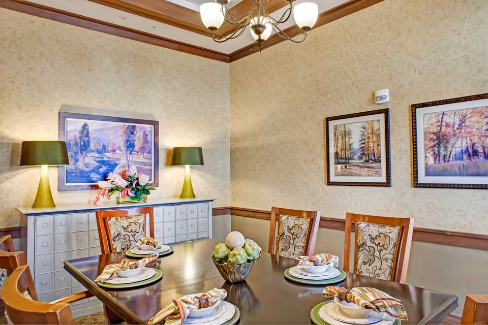 A dining room with a dark wooden table set for six, featuring plates, bowls, and striped napkins with napkin rings. The chairs have patterned upholstery. On the wall behind the table are three framed landscape paintings. A sideboard with two green lamps and a floral arrangement is also visible. The room has beige textured walls with wooden trim and a chandelier overhead.
