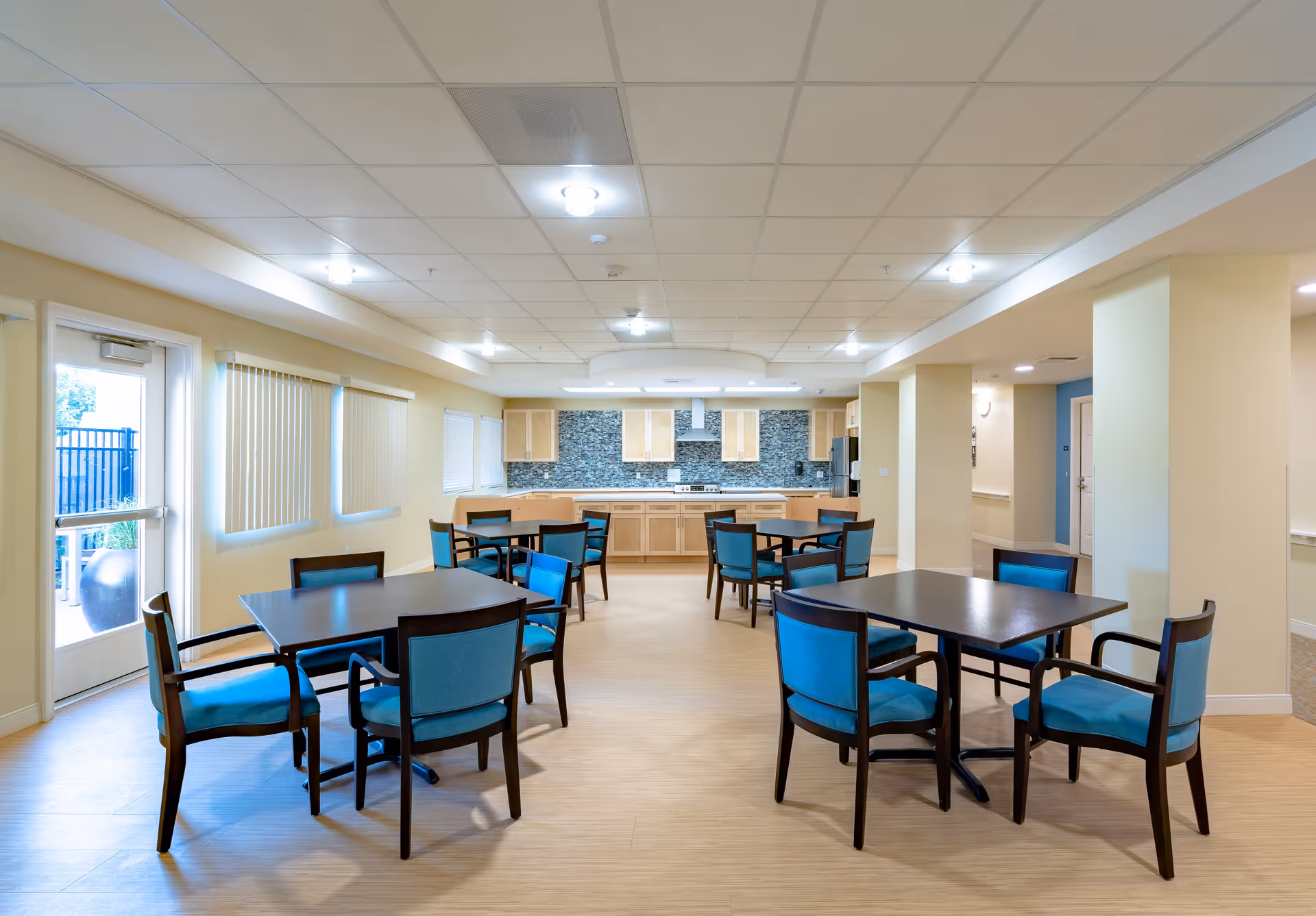A bright and clean dining area with several dark wooden tables and blue cushioned chairs arranged neatly. The room has light-colored walls and a light wood floor. At the back, there is a kitchen area with light wood cabinets, a stove, a refrigerator, and a tiled backsplash. Large windows with vertical blinds and a glass door letting in natural light are on the left side.