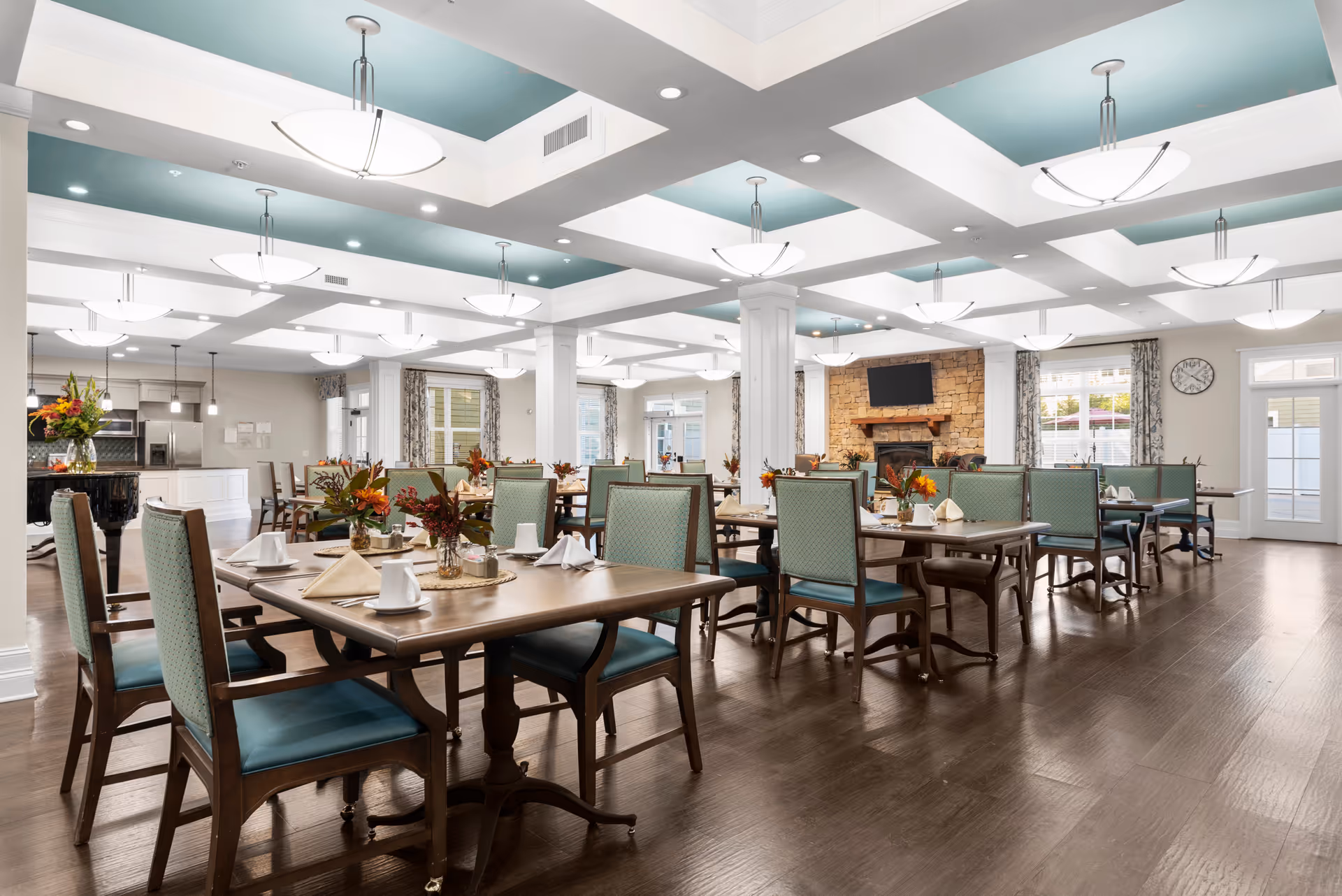 A spacious dining room with multiple wooden tables and green cushioned chairs. Each table is set with white cups, napkins, and small floral centerpieces. The room features a coffered ceiling with teal accents and modern light fixtures. There is a stone fireplace with a mounted flat-screen TV above it, and large windows with patterned curtains allowing natural light to enter.