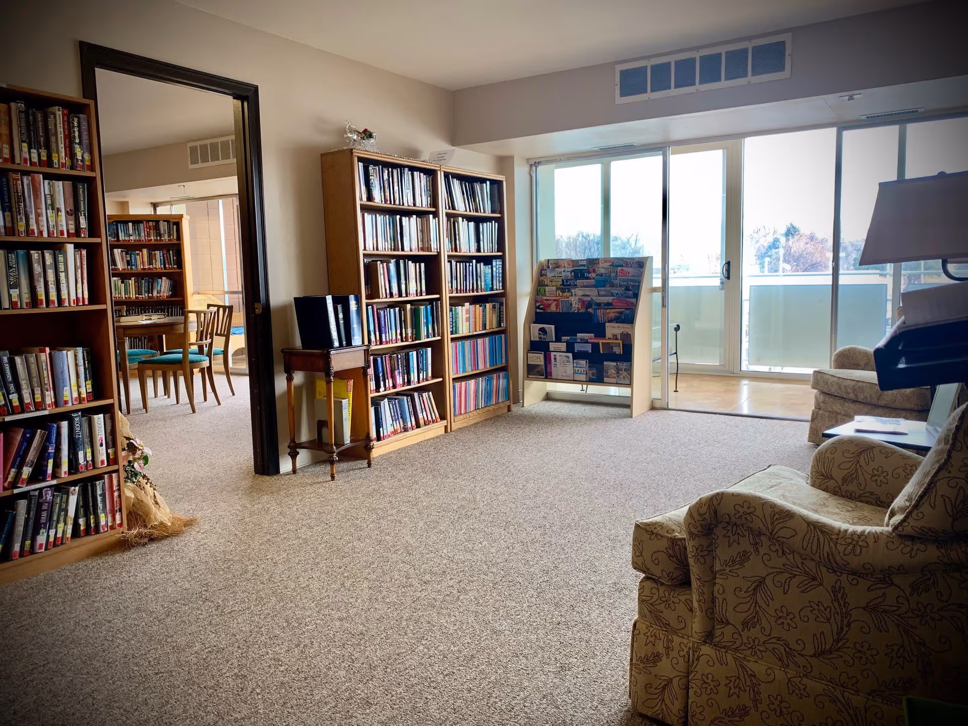 A cozy library or reading room in an assisted living center with bookshelves filled with books, a magazine rack, comfortable armchairs, and large sliding glass doors leading to a balcony with a view outside.