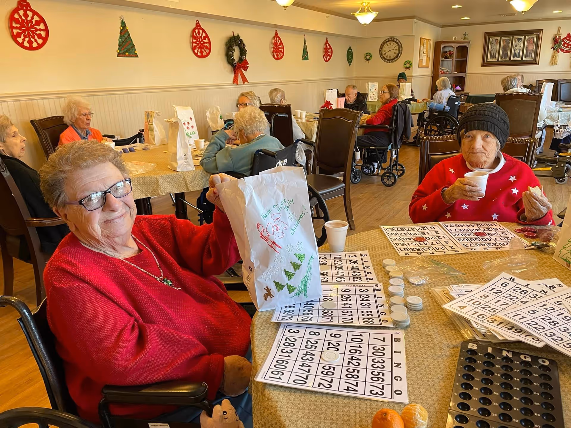 Elderly residents in a decorated common room playing bingo at tables, holding snacks and drinks.