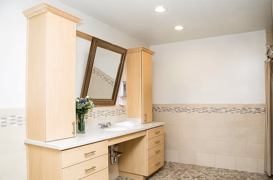 A clean bathroom vanity area with light wood cabinets, a white countertop with a built-in sink, a tilted rectangular mirror, a vase with flowers, and a soap dispenser mounted on the wall. The walls are tiled halfway up with a decorative mosaic border, and the floor has small square tiles in neutral tones.