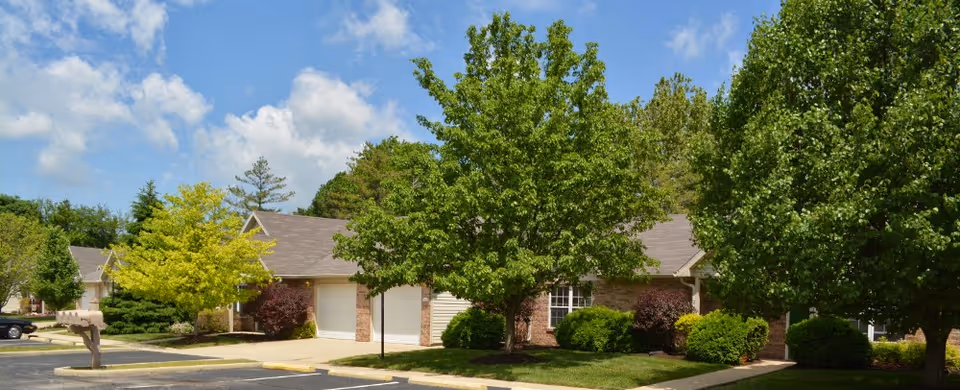 Exterior view of a residential building with a brick and siding facade, surrounded by green trees and shrubs under a partly cloudy blue sky. A parking lot and mailbox are visible in the foreground.