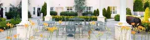 Outdoor courtyard with metal tables and chairs, blooming flowers near a columned building.