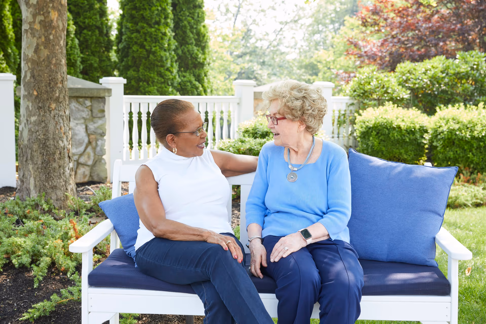 Two elderly women sitting on a white outdoor bench with blue cushions, engaged in conversation in a garden setting with green shrubs, trees, and a white fence in the background.