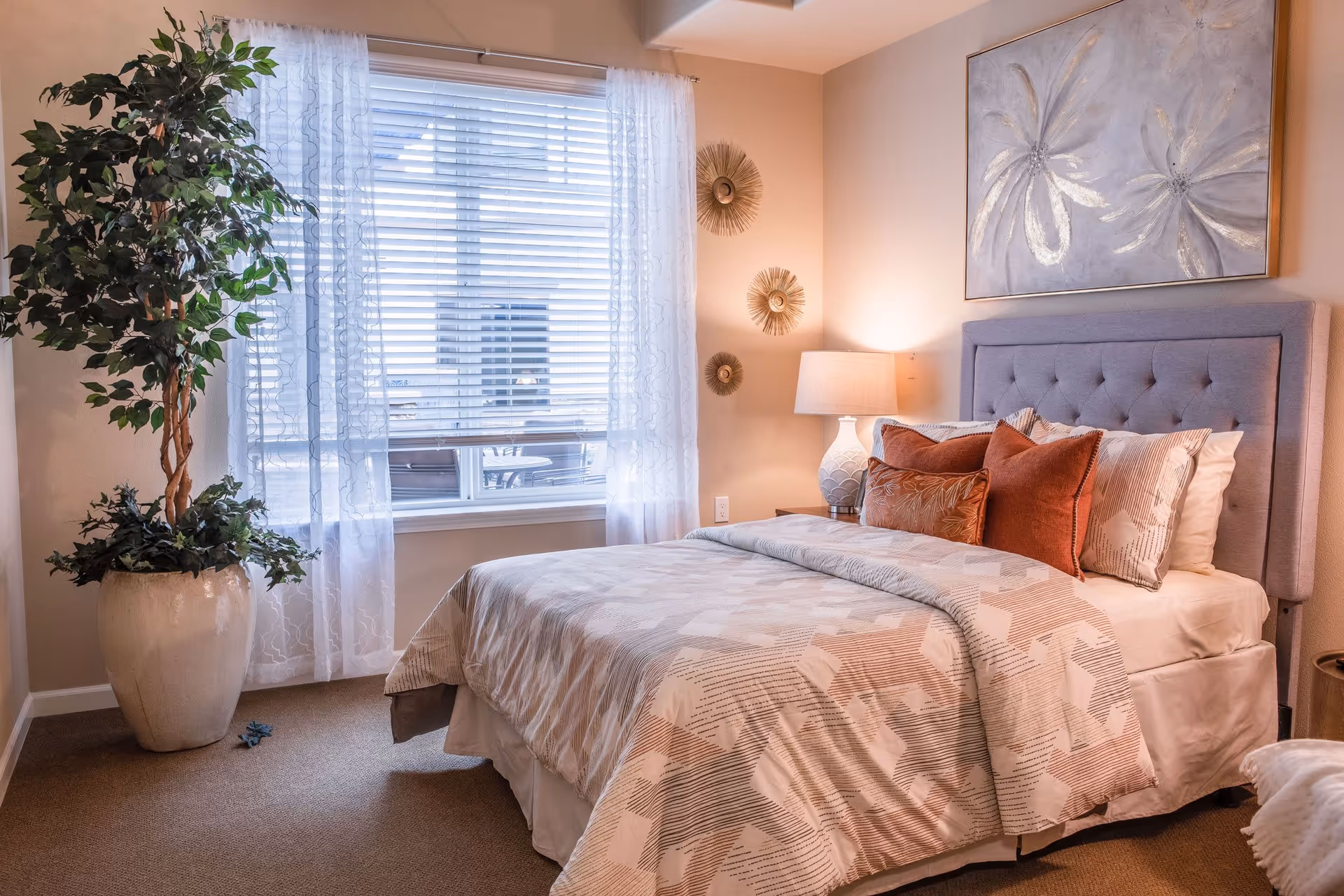 A cozy bedroom with a neatly made bed featuring a patterned beige and white comforter and several decorative pillows in shades of rust and beige. A tufted gray headboard is against the wall, which is adorned with a large floral painting and three round decorative wall hangings. To the right of the bed is a nightstand with a white lamp. A large window with white blinds and sheer curtains lets in natural light. A tall potted plant is placed in the corner near the window.