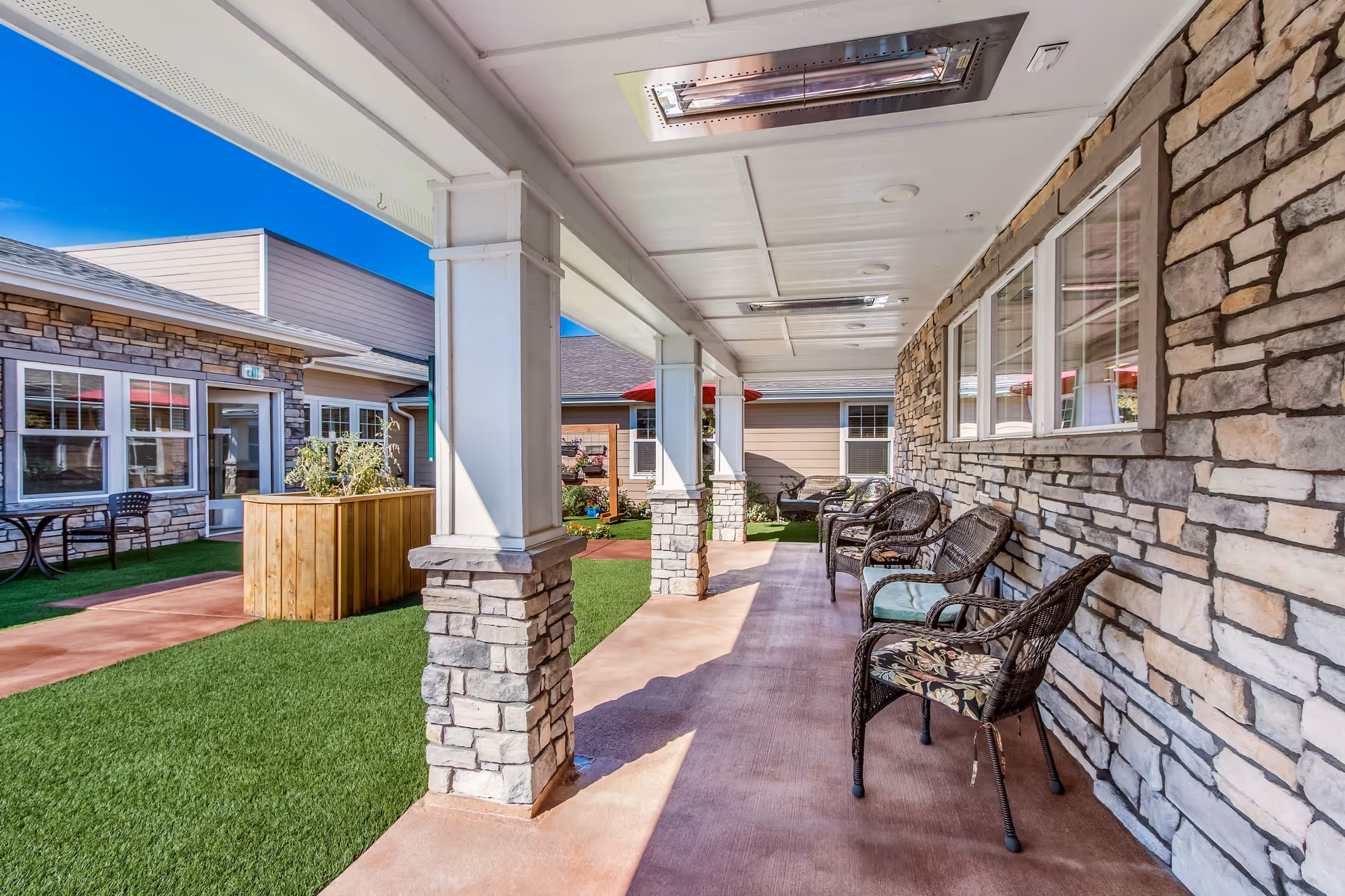 Covered outdoor patio area with stone pillars and wicker chairs with cushions along a stone wall. The patio overlooks a courtyard with artificial grass, a wooden planter box, and a table with chairs near the building. The sky is clear and blue.