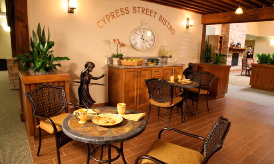 Indoor bistro seating area with round tables and chairs, plants, and a wall sign reading "Cypress Street Bistro".