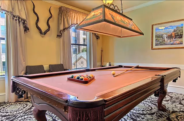 A room with a pool table set up for a game, featuring a rack of billiard balls and two pool cues resting on the table. The room has large windows with curtains, two chairs against the wall, decorative wall-mounted antelope horns, a hanging stained glass light fixture above the table, and a framed painting of horses on the wall.