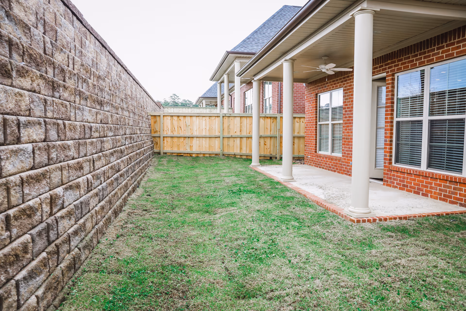 A small grassy backyard with a covered concrete patio supported by columns beside a brick house and a tall stone retaining wall.