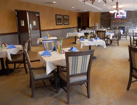 A dining room with multiple tables covered in white tablecloths, each set with blue folded napkins and small centerpieces. The room has beige walls, carpeted floors, wooden chairs with striped cushions, and a television mounted on a stone accent wall.