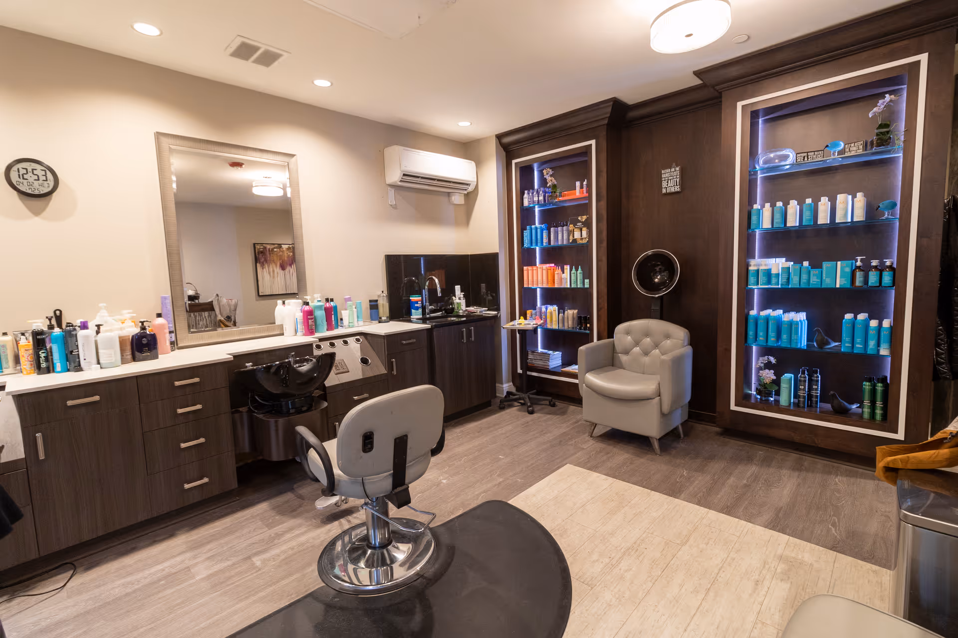 A well-lit salon room with styling chairs, a wash station, and shelves stocked with hair products.