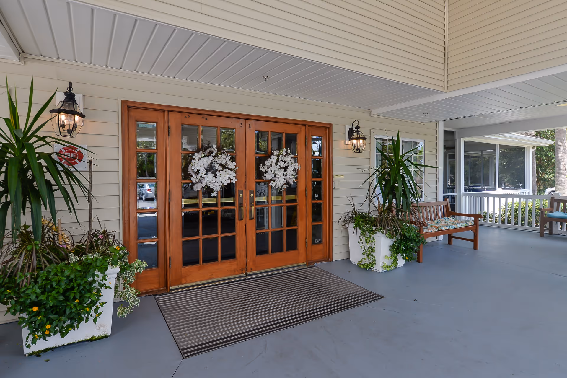Covered entrance area of a facility with double wooden doors decorated with white floral wreaths, flanked by large potted plants and wall-mounted lantern lights. There is a wooden bench with a floral cushion on the right side and a gray floor mat in front of the doors.