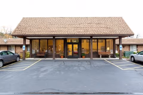 Front exterior view of Crystal Pines Rehabilitation & Health Care Center showing a single-story building with a peaked roof, large glass entrance doors, benches on either side of the entrance, potted plants, and parking spaces in front.