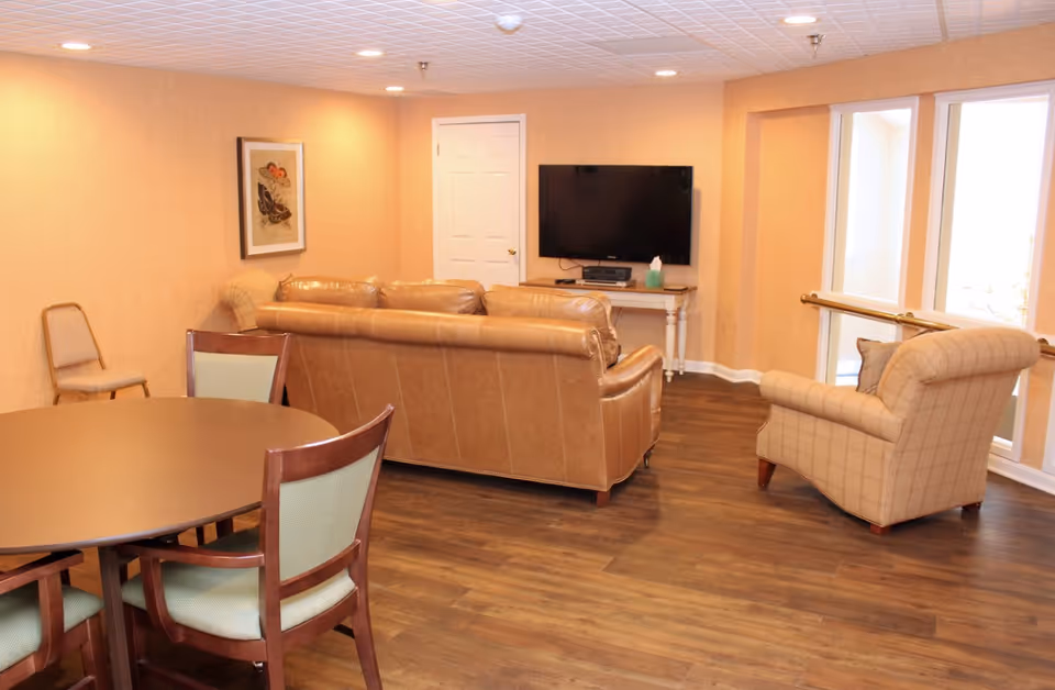 A cozy living room area with a round wooden table and four chairs in the foreground. Behind the table, there is a brown leather sofa facing a wall-mounted flat-screen TV above a small white table. To the right, there is a beige armchair with a pillow near large windows letting in natural light. The room has wood flooring and peach-colored walls with a framed picture on one wall.