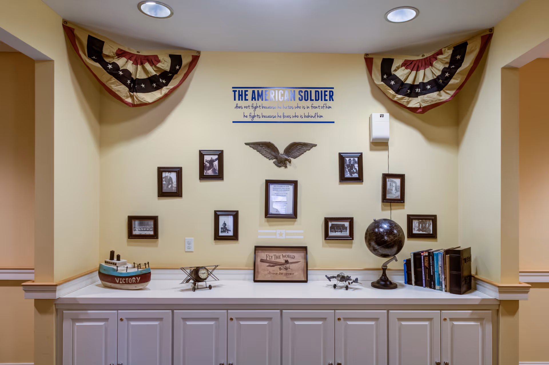 A patriotic-themed interior display above a white cabinet featuring framed photos, a globe, books, model boat and a sign reading 'The American Soldier'.