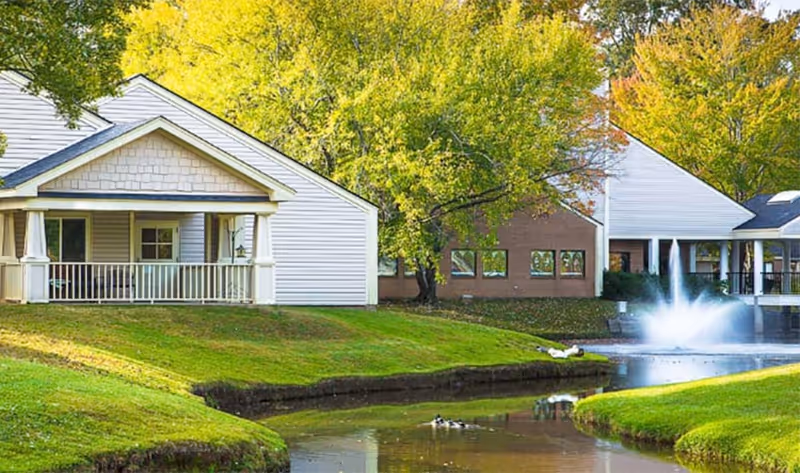 A peaceful outdoor scene at The Stiles Apartments featuring white residential buildings with porches, green grassy banks, a small pond with ducks, and a water fountain. Trees with green and autumn-colored leaves surround the area.