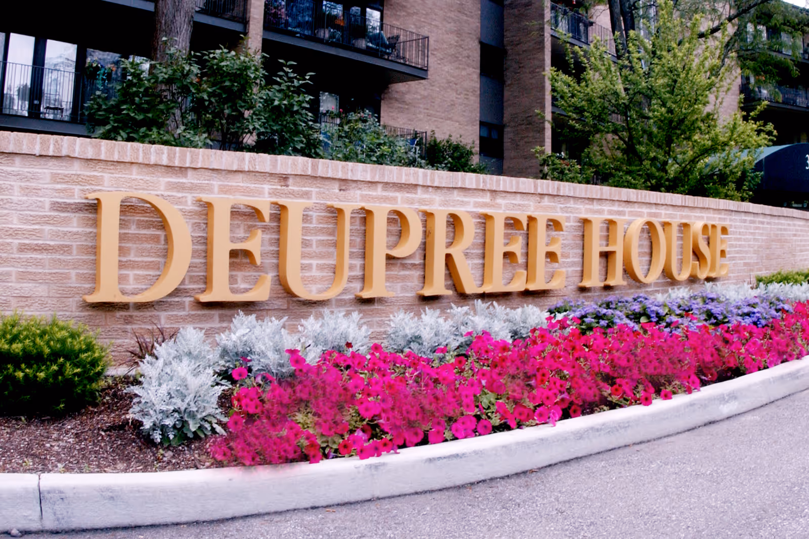 A brick wall with large gold letters spelling 'DEUPREE HOUSE' surrounded by colorful flowers and greenery, with a multi-story building in the background.