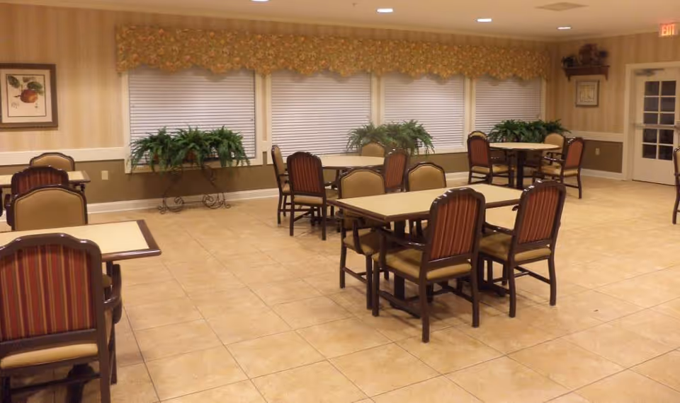 A dining room with several tables and chairs arranged neatly on a tiled floor. The walls have a beige striped wallpaper with a chair rail, and there are large windows with closed blinds and floral valances. Green potted plants are placed on stands in front of the windows. A framed picture hangs on the left wall, and a door with glass panes is visible on the right side.