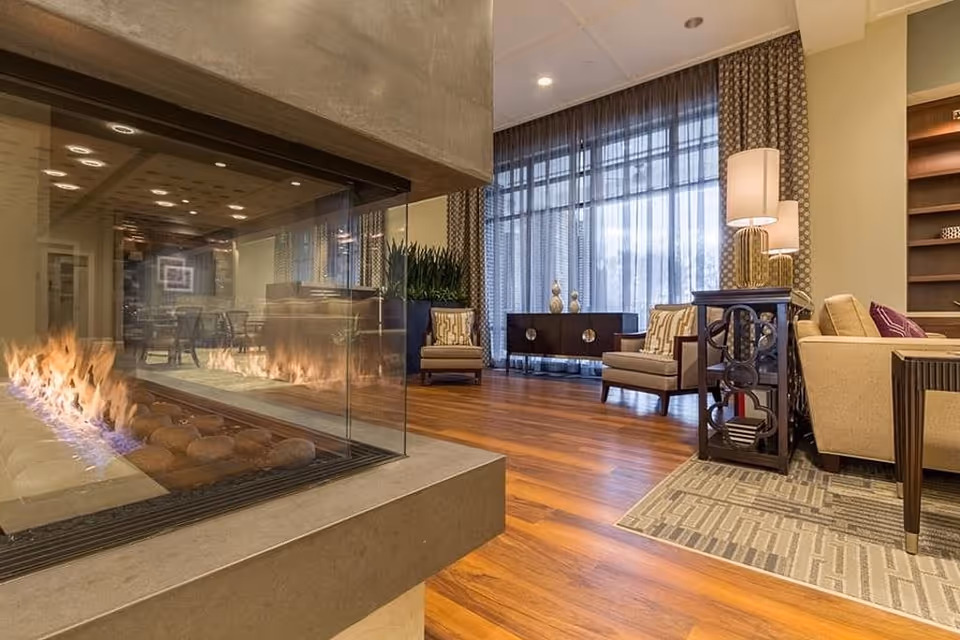 A cozy living room area with a modern glass-enclosed fireplace in the foreground. The room features wooden flooring, comfortable armchairs with patterned cushions, a side table with a lamp, a large window with sheer curtains, and a wooden cabinet against the wall.