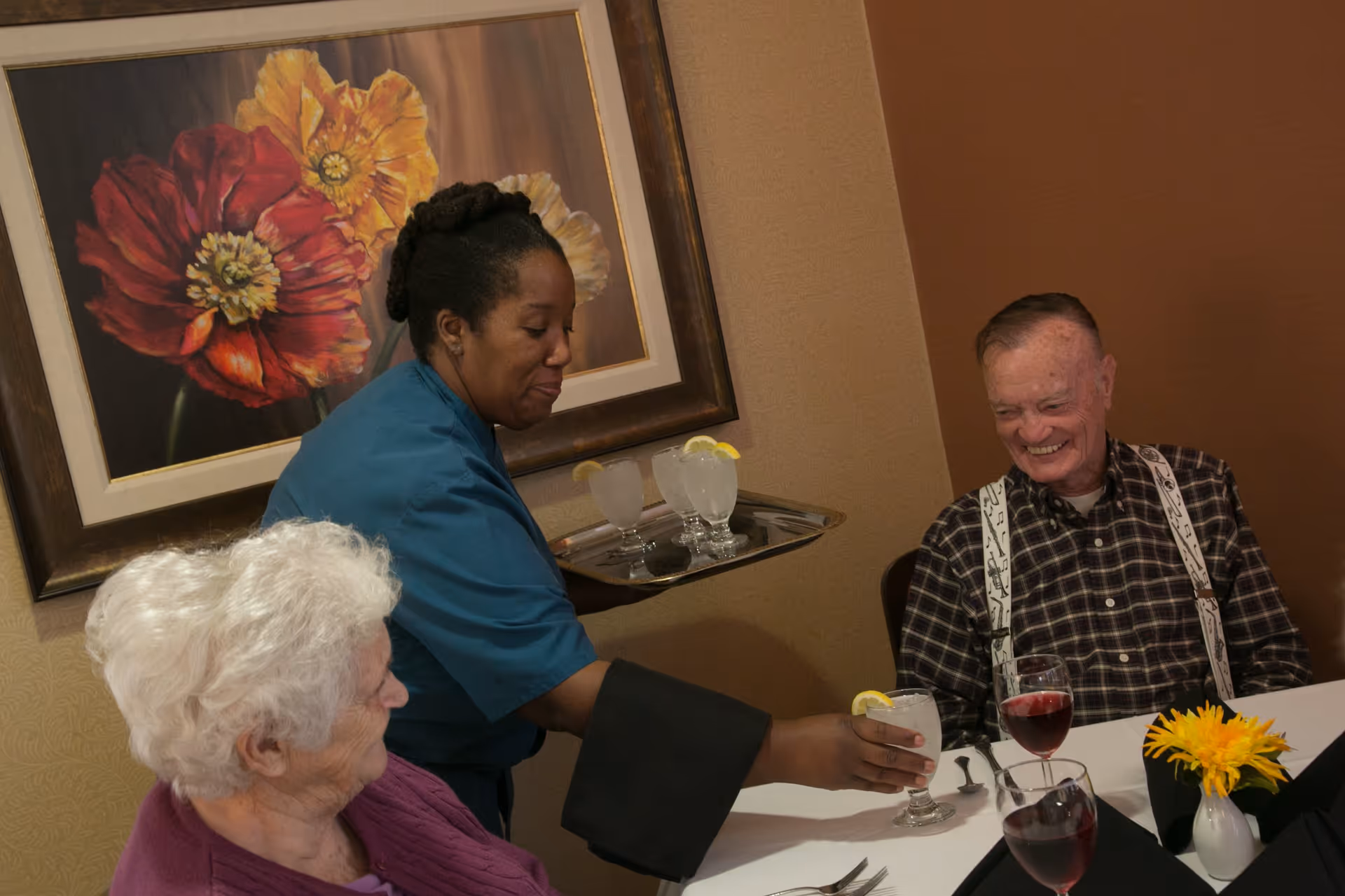 A caregiver in a blue uniform serving a glass of water with a lemon slice to an elderly man sitting at a dining table. An elderly woman with white hair is also seated at the table. The table is set with glasses of red wine, black napkins, and a small vase with a yellow flower. A large framed painting of red and yellow flowers hangs on the wall behind them.