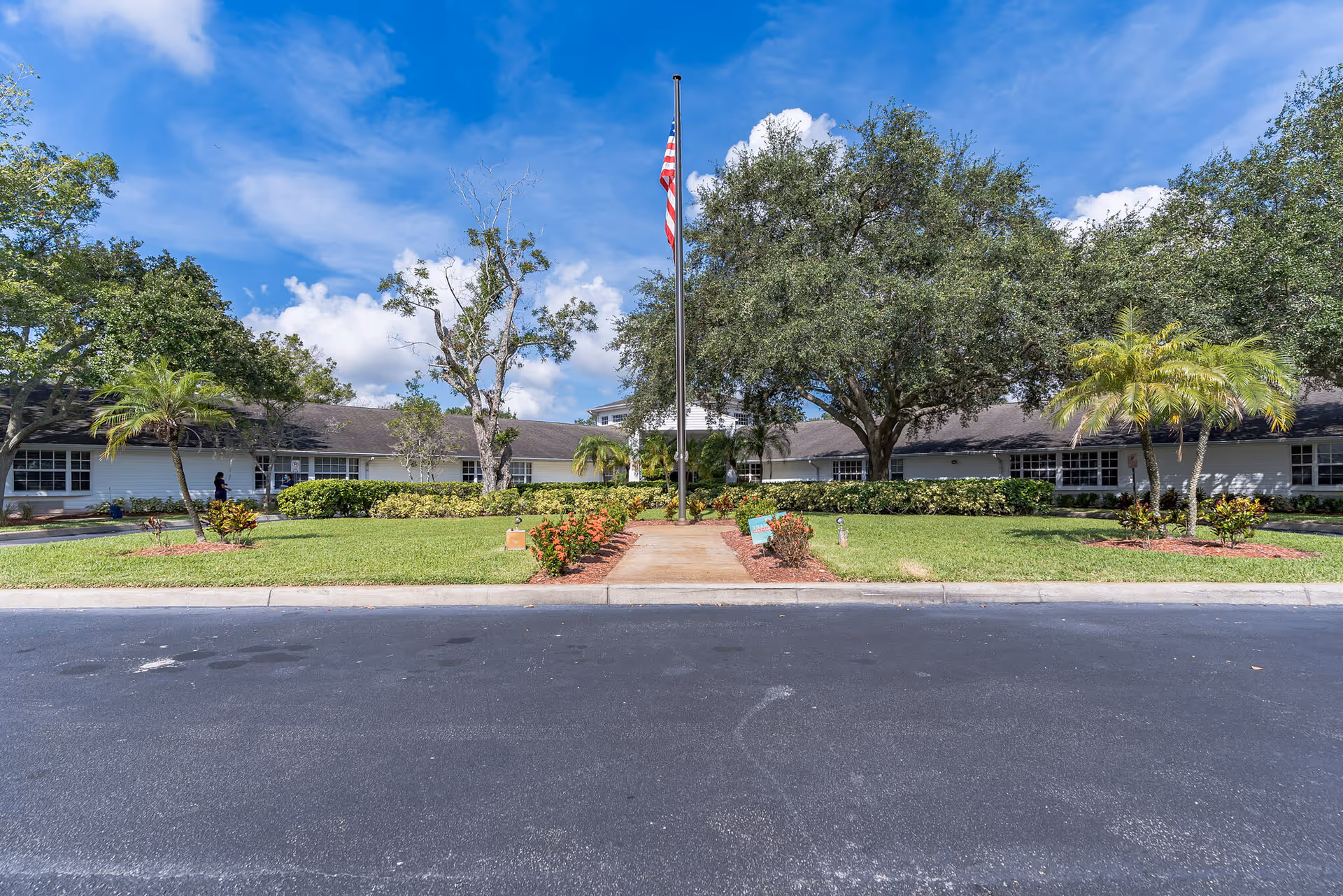 Exterior view of a single-story building with a well-maintained lawn, trees, and an American flag on a flagpole in the center. The sky is blue with some clouds.