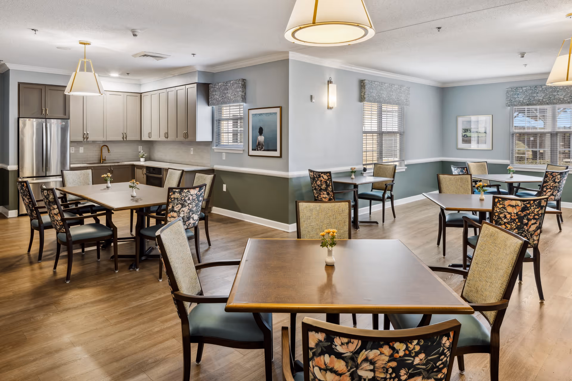 A bright and spacious dining area with multiple square wooden tables and chairs with floral and plain upholstery. The room features light blue and green walls with white trim, large windows with valances, and modern pendant lights. In the background, there is a kitchen area with gray cabinets, a stainless steel refrigerator, and a dishwasher.