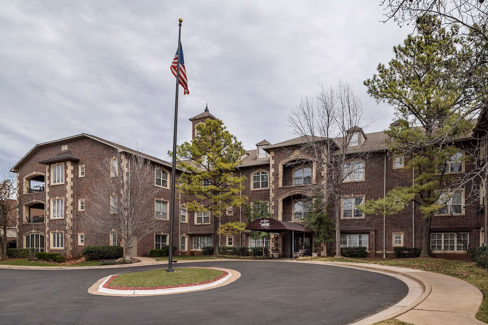 Exterior view of a three-story brick assisted living facility with a circular driveway and an American flag on a flagpole in front. The building has multiple windows and a covered entrance with the name 'The Mansion at Waterford Assisted Living' visible above the entrance. Trees and shrubs surround the building.