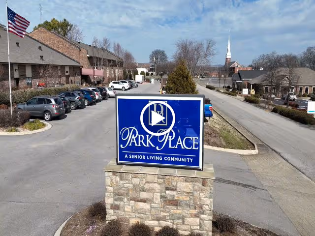 Blue stone monument sign reading 'Park Place, A Senior Living Community' at the facility entrance with parked cars, buildings, and a road in the background.