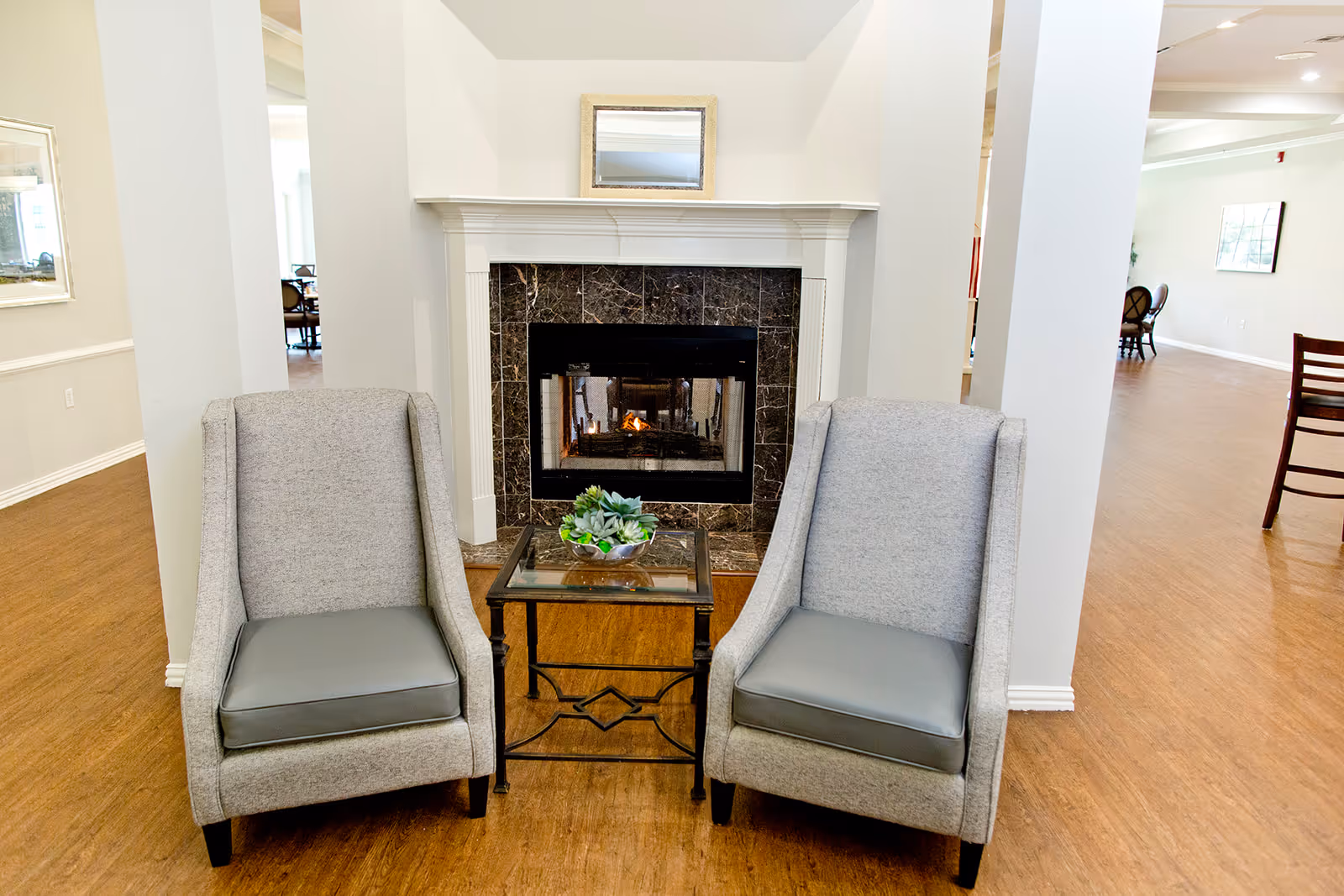Two gray armchairs with dark gray seat cushions are positioned on either side of a small glass-top table with a decorative plant on it, in front of a fireplace with a dark marble surround and a small mirror above the mantel in a spacious room with wood flooring.