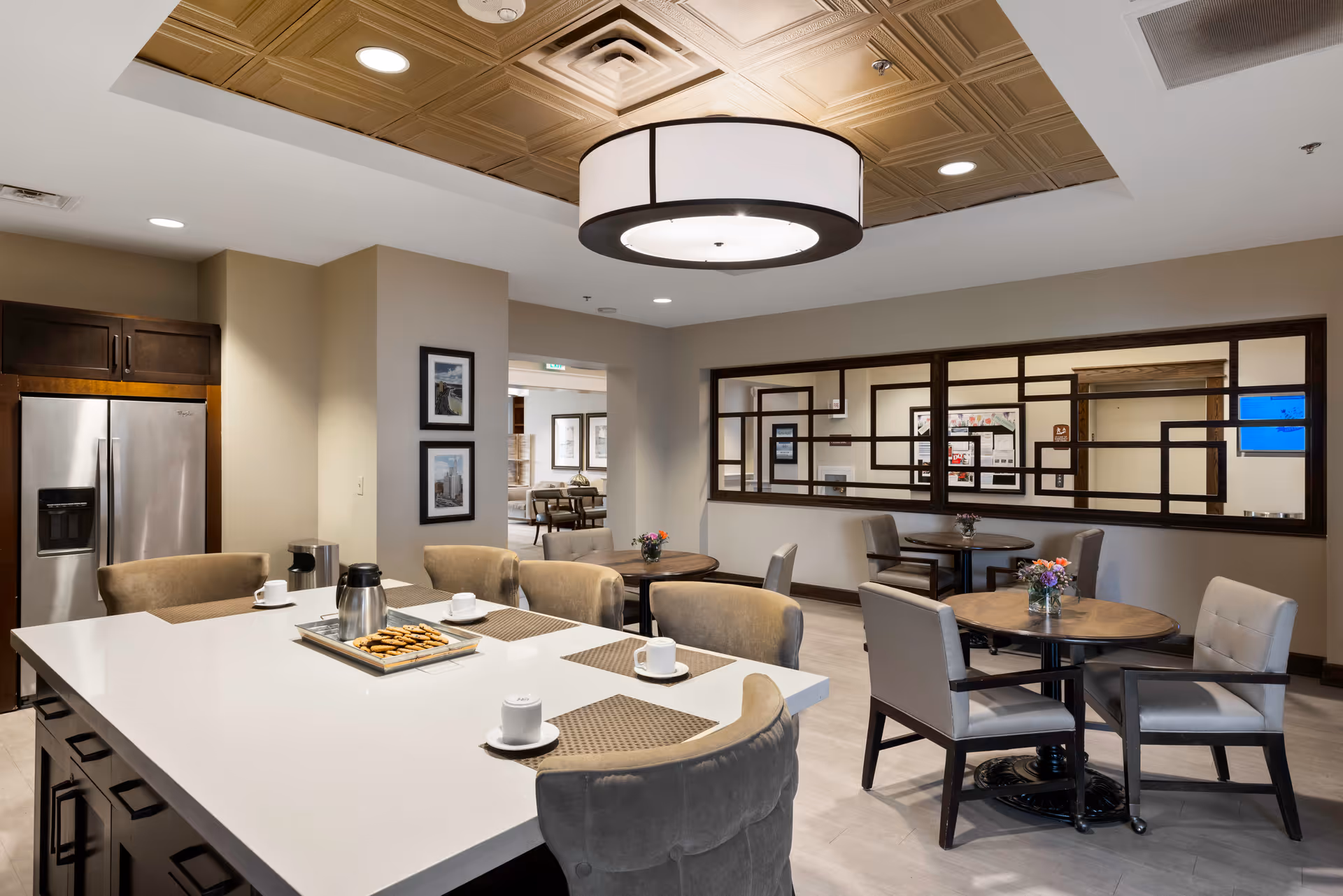 Modern communal dining and kitchen area with a large island, round tables, chairs, and a decorative ceiling light.