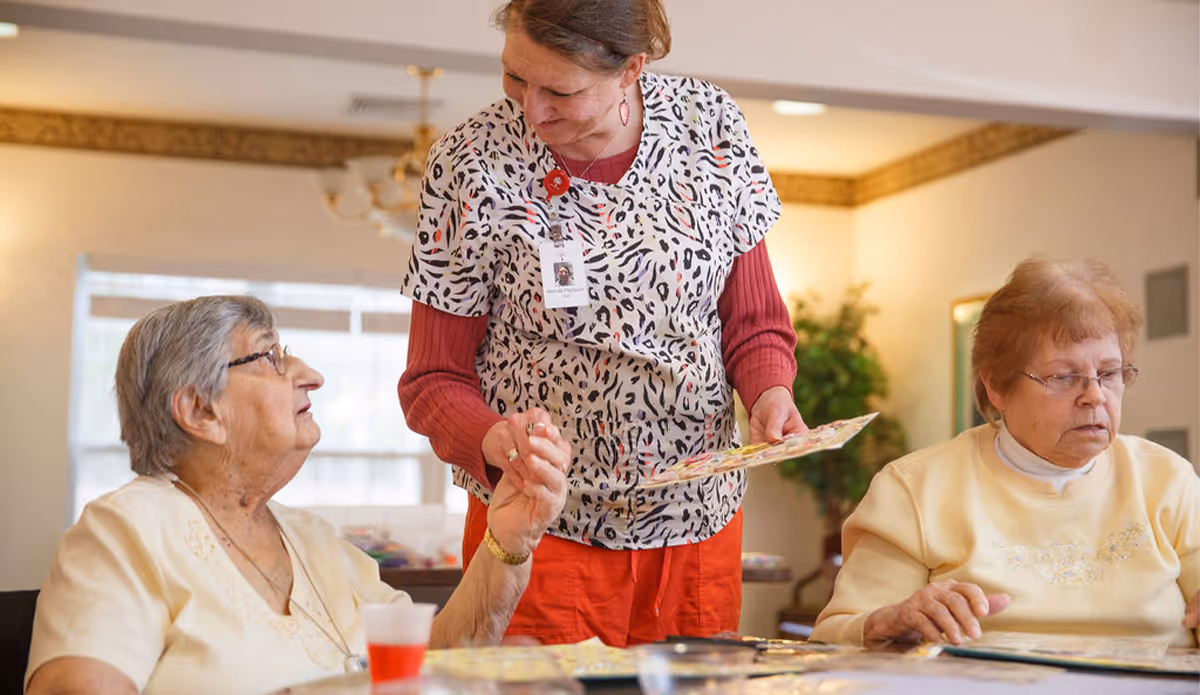 A caregiver in a patterned scrub top and red pants interacts with two elderly women seated at a table, engaging in an activity with colorful cards in a well-lit room with a window and decorative molding.