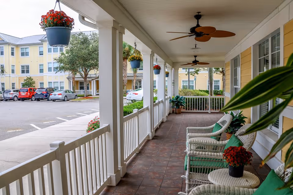 Covered outdoor porch area with white wicker chairs and green cushions, small round table with a potted red flower, ceiling fans, hanging flower pots, and a view of the parking lot and yellow senior living building in the background.